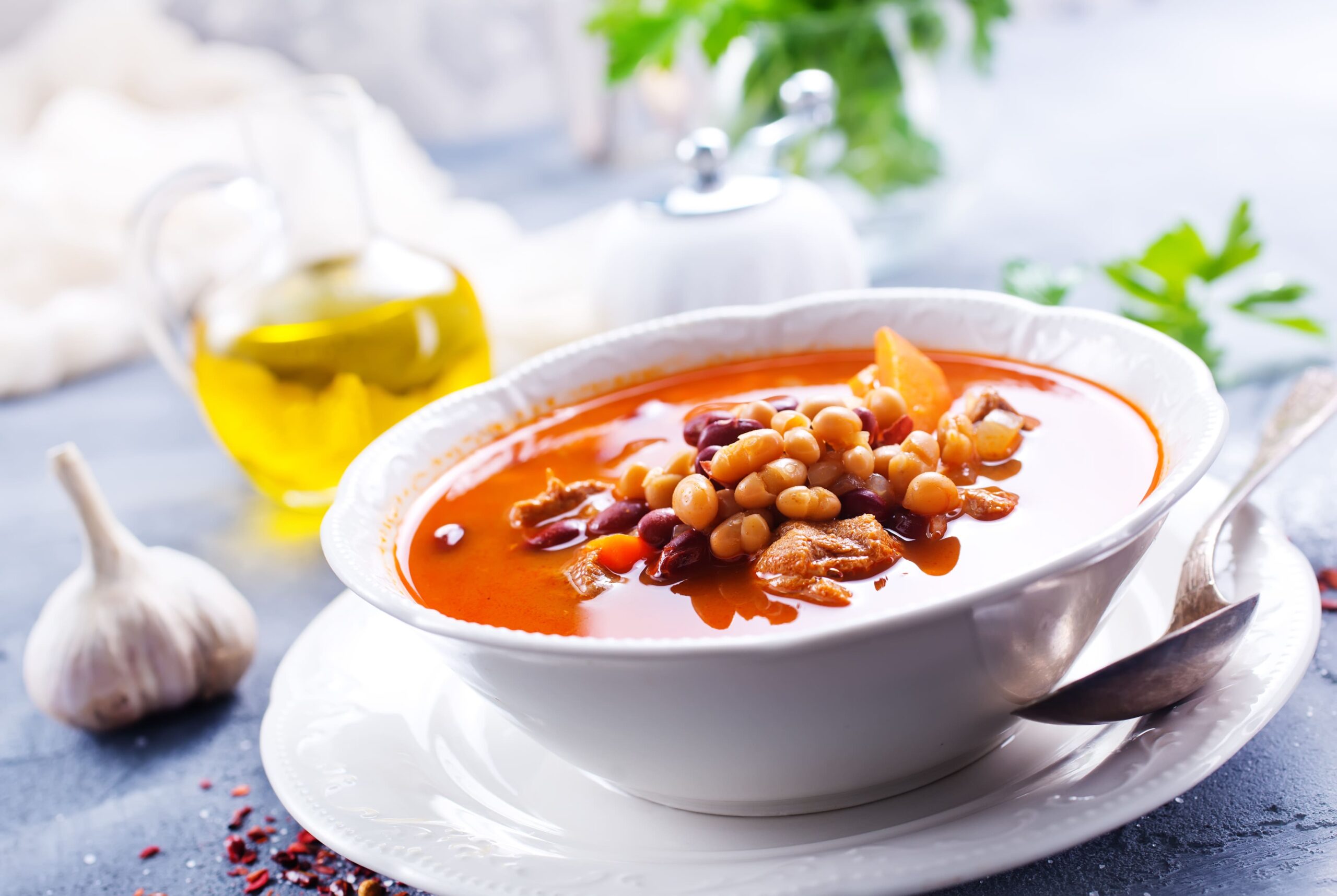 Fasolada, sopa griega de alubias, servida en un plato hondo, con verduras y hierbas. Al fondo, una botella de aceite de oliva, una cabeza de ajo y hojas verdas decorativas. 