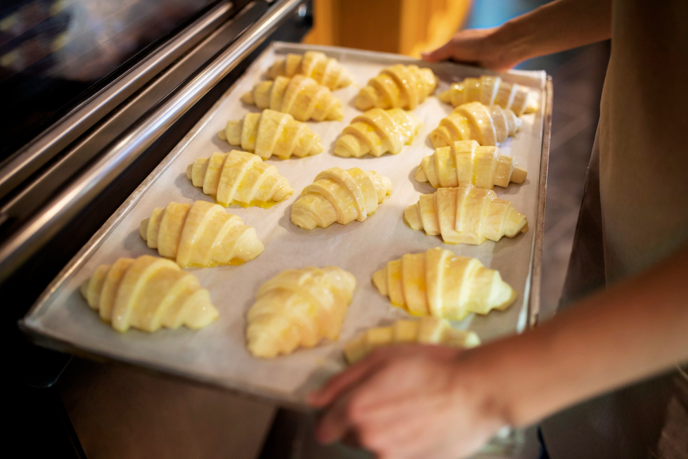 Una persona lleva entre las manos una bandeja con varios croissant dispuestos ordenadamente sobre papel encerado, listos para meter al horno.