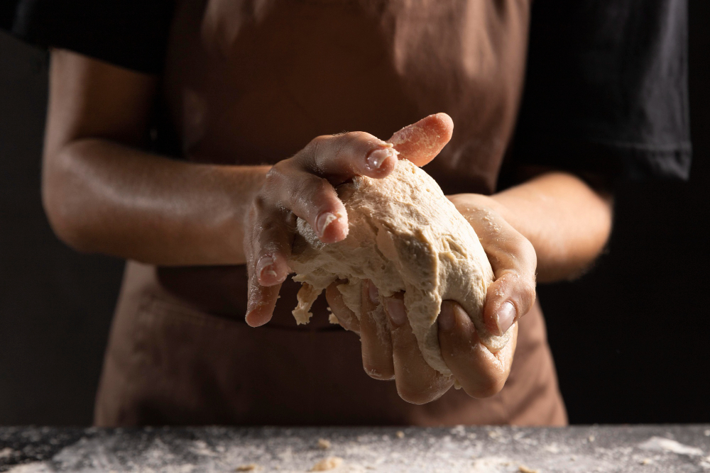 Vista frontal de una persona preparando la masa para el pan. El corte de la imagen est{a hecho de la cadera hacia los hombros, de tal forma que solo se muestran las manos amasando.