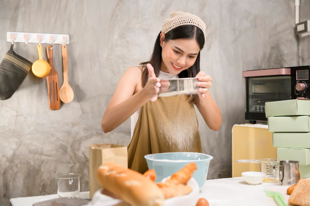 Mujer joven sonríe en su cocina mientras cierne harina en bowl. La rodean utencilios de cocina, variedad de panes y al fondo un pqueño horno convencional. Su delantal está salpicado de harina.