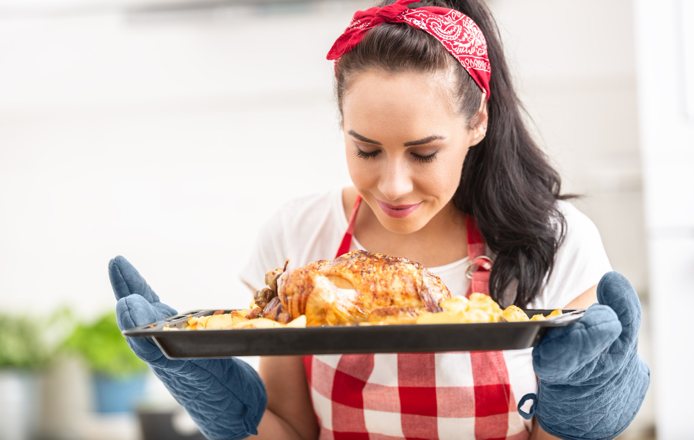 Mujer joven disfruta del aroma de un pollo recién horneado mientras lo sostiene con cuidado sobre una bandeja. Lleva puestos guantes de cocina y un delantal a cuadros. 