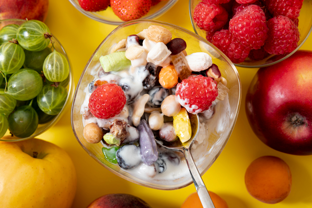 vista cenital de ensalada de frutas con crema y leche condensada, presentada en un recipiente de cristal. A su alrededor, se aprecian frutas frescas como manzanas, uvas y fresas.