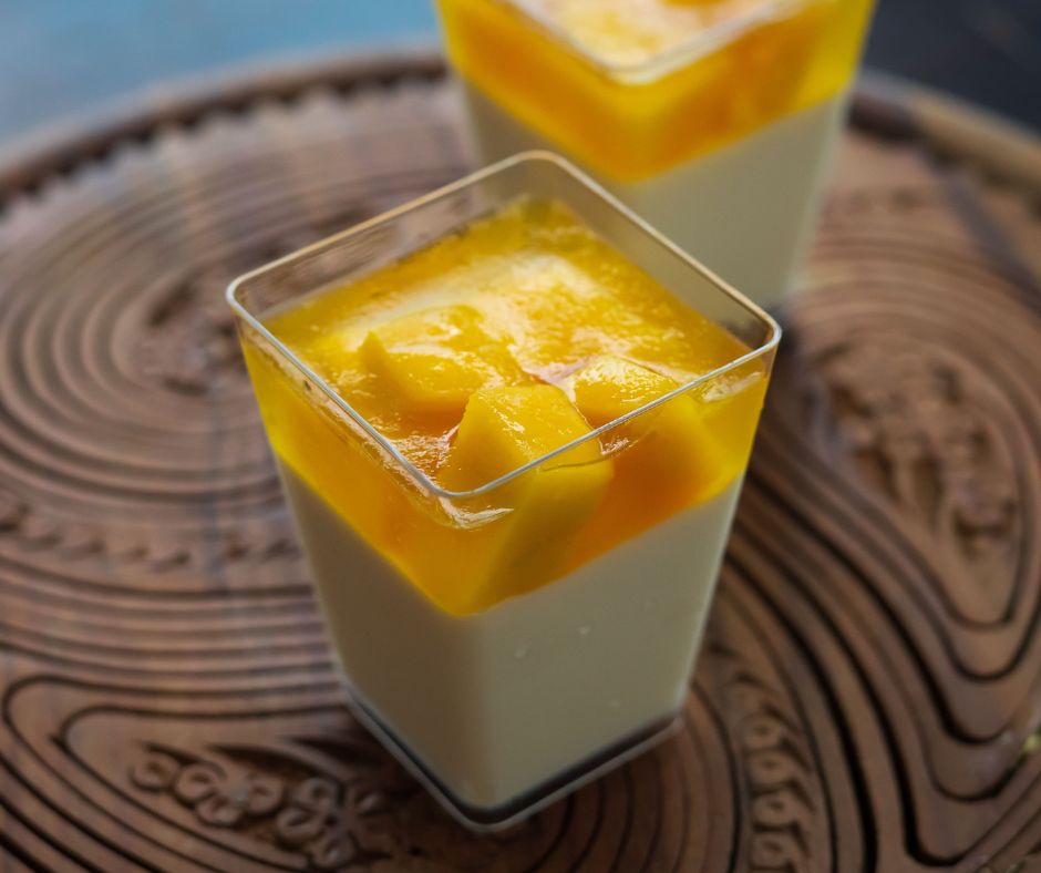 2 vasos cuadrados de plástico transparente con gelatina de queso en la parte inferior, y mango, en la parte superior, decorados trozos de la misma fruta.