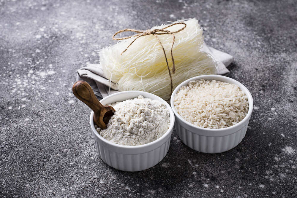 Fideos de arroz crudos, envueltos finamente con un lacito, dispuestos junto a dos recipientes pequeños, un con harina, el otro con granos de arroz.