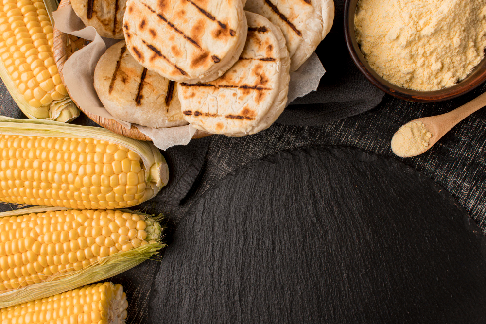 Vista cenital de arepas caseras, acompañadas de mazorcas de maíz y un pequeño recipiente con harina junto a una cucharilla de madera.