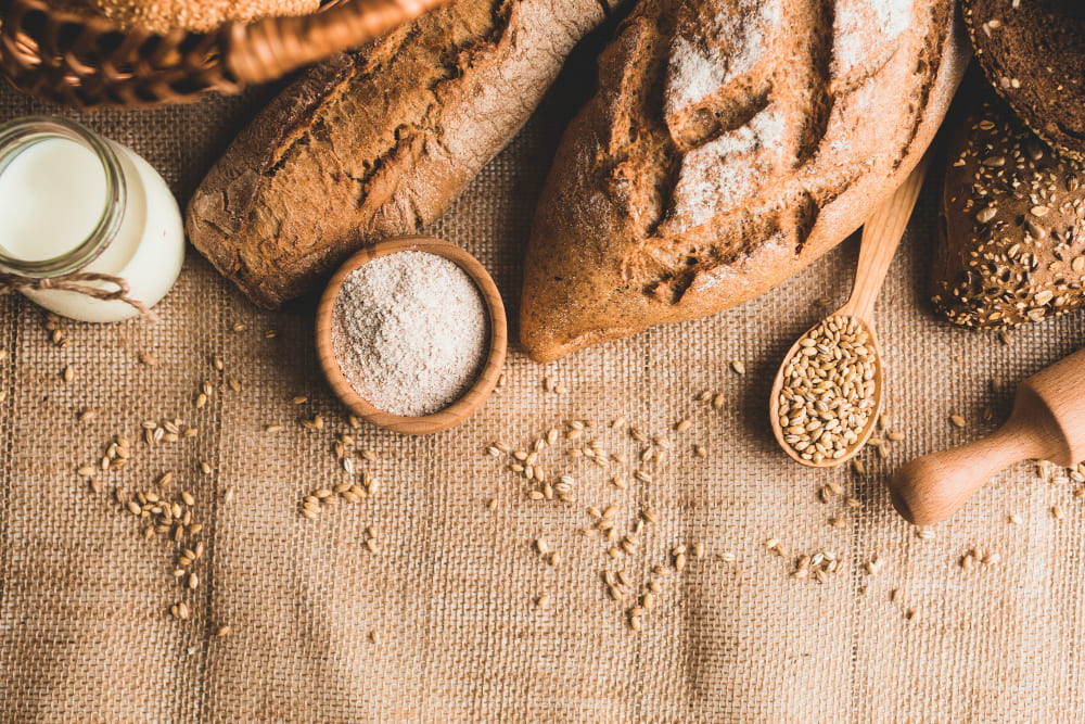 Vista cenital de panes junto a pequeños recipientes con harina y granos esparcidos alrededor. 