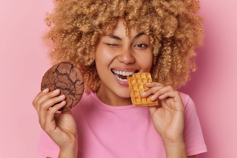 Mujer joven con pelo rizado sonr{ie mientras giña un ojo y muerde un waffle que sostiene con una mano. En la otra mano sostiene una galleta de chocolate.