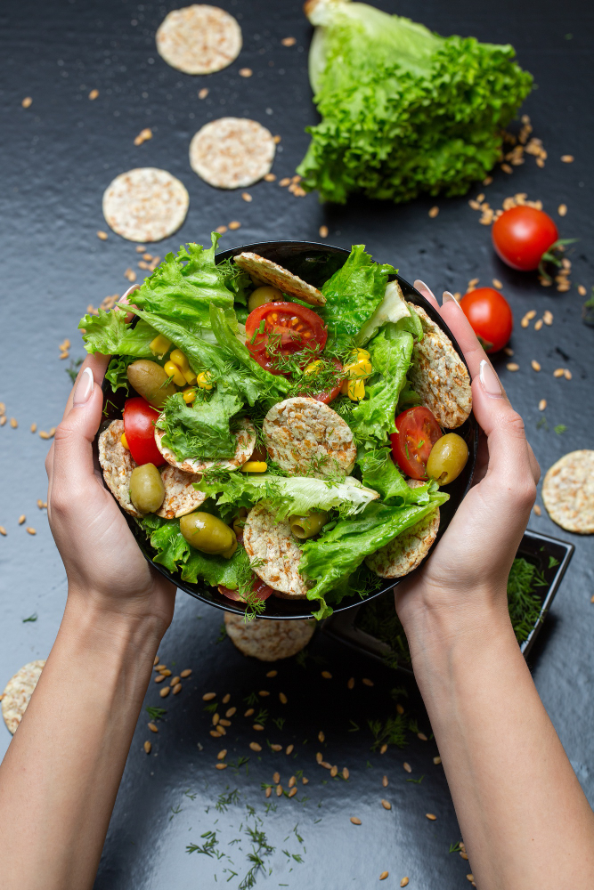 Vista cenital de dos manos sosteniendo un bowl con una ensalada mediterranea, fresca, colorida, con vegetales, granos y galletas saladas.