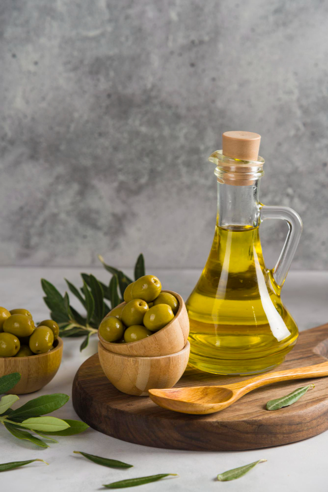 Botella de aceite de oliva con tapón de corcho sobre tabla de madera. La esena está acompañada por recipientes y una cuchara, también de bambú y aceitunas frescas.
