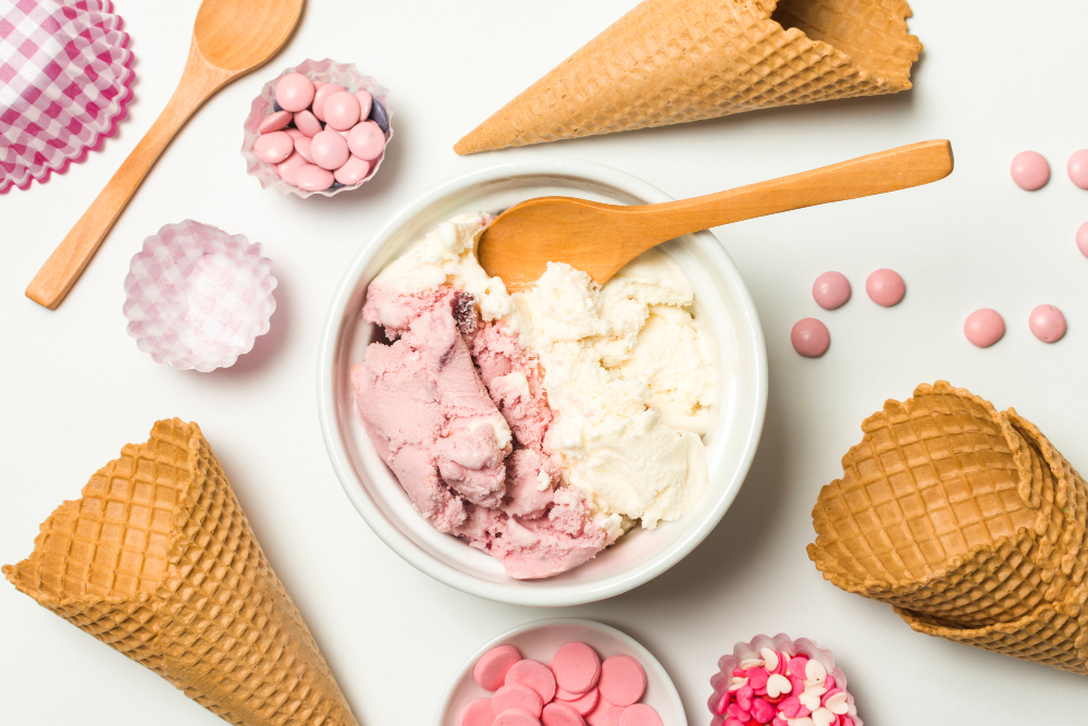 Vista cenital de un plato con helado de dos sabores: fresa y crema, tiene una cuchara de bambú hundida en medio. La escena está decorada también con confituras rosas, conos de galleta y capacillos para postre.