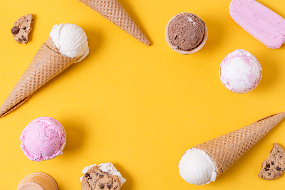 vista cenital de variedad de helados cremosos servidos en conos y vasitos sobre un fondo amarillo vibrante. Algunos de los helados están acompañados con galletas de chispas de chocolate.