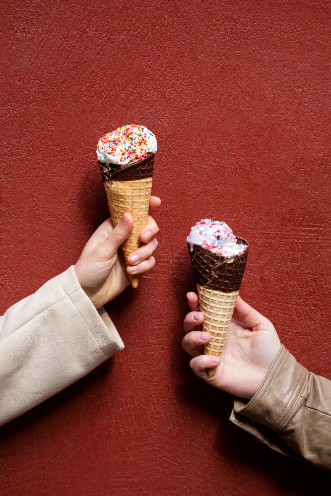 Dos personas disfrutando helados. En la imagen únicamente se muestran los brazos y las manos sosteniendo conos con cobertura de chocolate que cxontienen helados de fresa con chispas dulces de colores. La imagen tiene tonos terrosos, destaca el fondo color chedrón.
