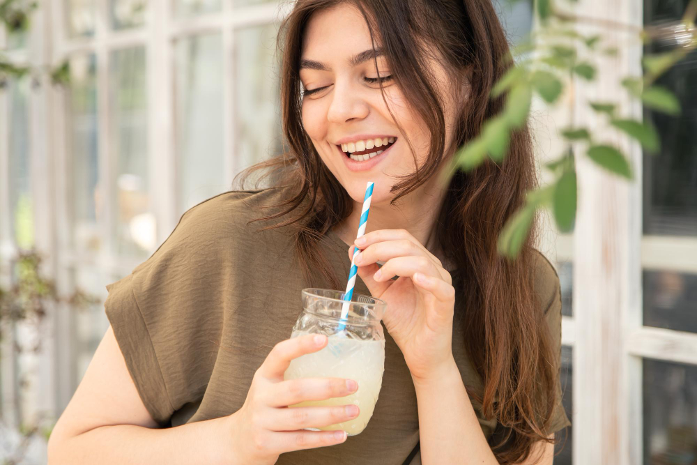 Una mujer joven de cabello castaño disfruta un vaso de refrescante agua de frutas, mientras sonríe.