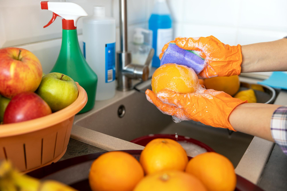 Una persona lava una naranja con agua y jabón, para preparar agua de frutas. 

La persona está frente a una tarja, lleva puestos unos guantes de látez de color naranja. Sobre el fregadero hay diferentes productos de limpieza, como jabones y desinfectantes. 

A un lado hay dos charolas con más frutas para ser lavadas.